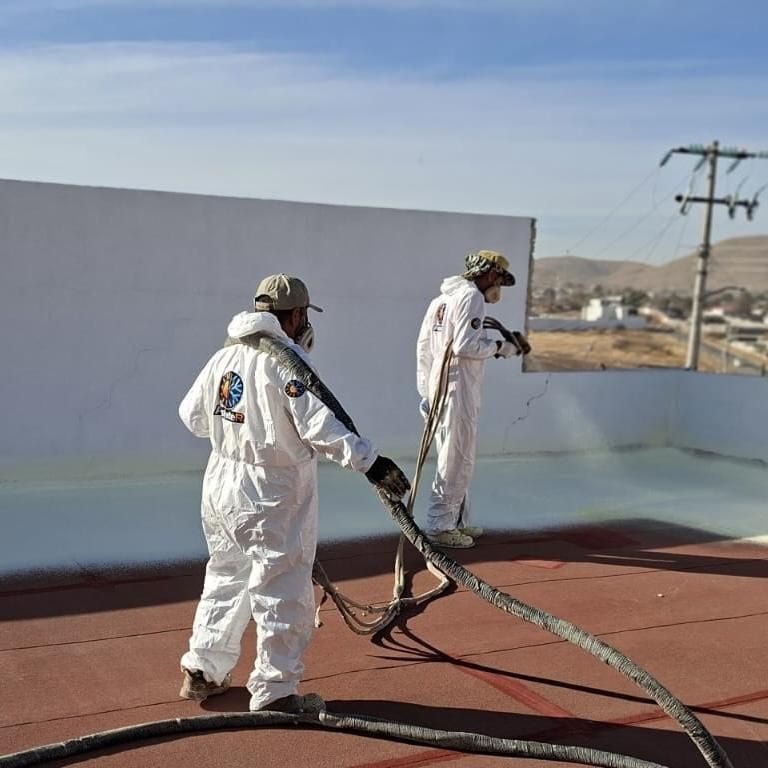 Two workers in white protective suits and respirators spray coating on a metal roof surface with industrial equipment visible.